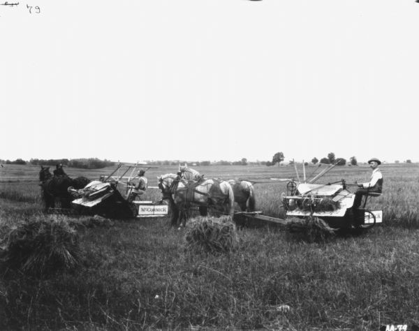 Two men are using horse-drawn McCormick binders in a field. There are sheaves of wheat piled in the foreground.