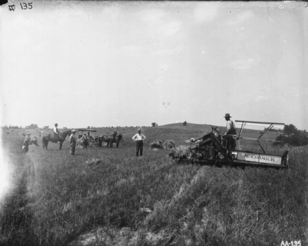 View from rear of a man using a horse-drawn McCormick binder in a sloped field on the right. On the left a group of people are posing. Men and a young boy are standing, another man is on horseback, and in the background two people are near a horse-drawn buggy.