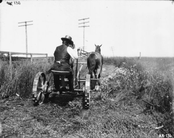 Rear view of a man using a horse to pull mower along a roadside. He is holding onto the brim of his hat. There is a wooden fence on the left, and three utility poles.