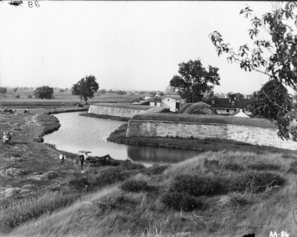 View down hill towards men standing near a horse-drawn carriage on the shoreline of a river. On the other side of the river is a high stone wall with grass growing on the top. Buildings are behind the wall on the right. A tall grain elevator is in the far background. Men are working in the field on the left.