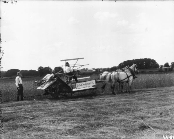 "Weisbrod farm." View of a man riding a horse-drawn McCormick binder on a transport truck in a field. A man is standing behind the binder on the left. Fields, trees and farm buildings are in the background.