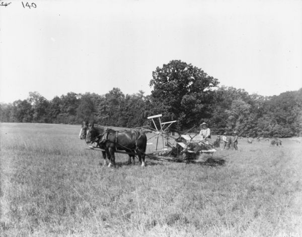 View across field towards a man using a horse-drawn McCormick binder in a field. Two men and a young boy are standing in the background holding sheaves of grain. Trees are at the edge of the field.