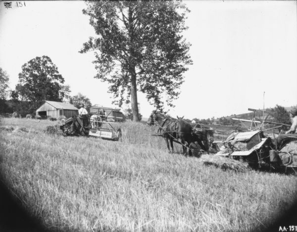 Two men using horse-drawn McCormick binders in a field on the slope of a hill. In the background are farm buildings, and trees and hills are in the far background.