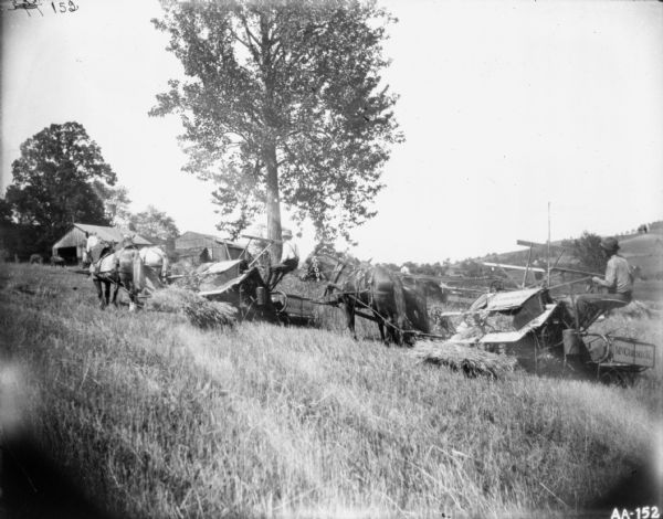 Two men using horse-drawn McCormick binders in a field on the slope of a hill. In the background are farm buildings, and trees and hills are in the far background.