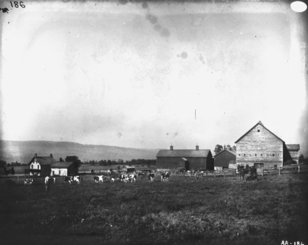 View across field towards cattle in a pasture. There is a farmhouse on the left, and farm buildings in the center. A farmer stands in the field on the left, and another man is using a horse-drawn mower on the right.