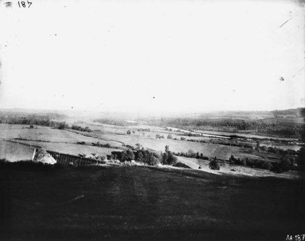 Landscape view down hill towards valley, with a railroad bridge over a river. Pasture lands are in the distance.