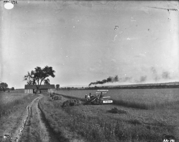 Landscape view of a man using a horse-drawn McCormick binder in a field. There is a barn near a tree further down the field. In the distance a railroad train is passing, and a long trail of smoke is coming from the chimney. A ridge of hills is in the far background.