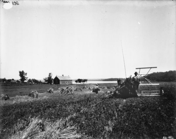 Rear view of a man using a horse-drawn McCormick binder in a field. A man is standing in the field holding sheaves of harvested grain. In the distance are farm buildings near a body of water.