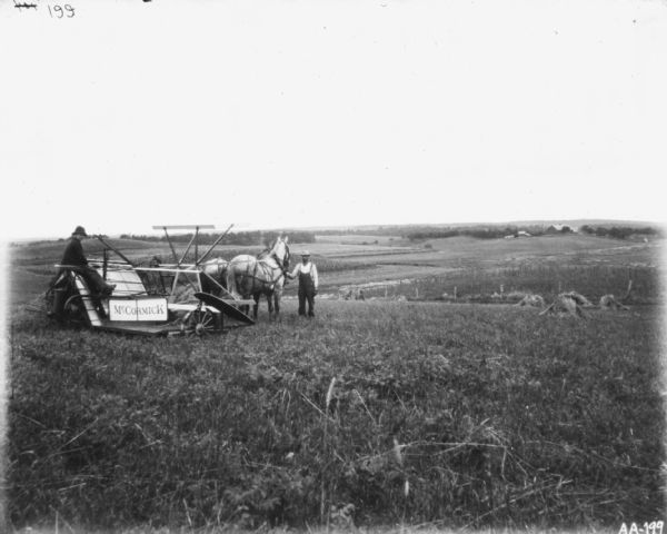 A man is standing and holding the horses of a horse-drawn McCormick binder in a field. Another man is sitting on the binder. In the background is a landscape view of fields and farm buildings.