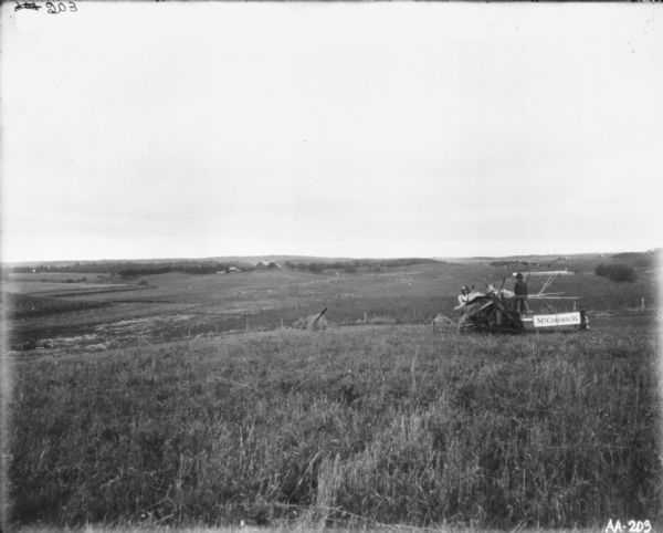 View across field towards a young man on a horse-drawn McCormick binder. A man is standing near the horses. Farm buildings are in the distance down a hill across fields.