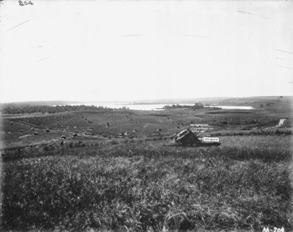 Landscape view of a McCormick binder in a field on a hill. Below are more hills with stacks of sheaves of grain. There is a river or lake in the background.