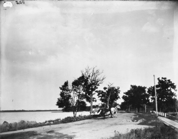 View of a man on a McCormick binder on a horse-drawn transport truck, on a road near a river or lake on the left. On the right is a board sidewalk.
