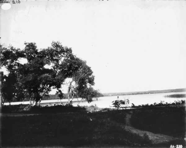 Man traveling on road near shoreline. He is riding on a McCormick binder on a horse-drawn transport truck. In the distance is the far shoreline.