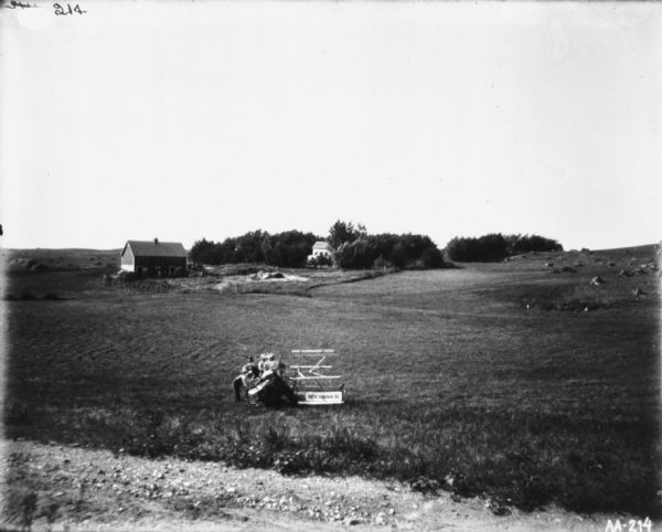 View down hill towards two men standing with a horse-drawn McCormick binder in a field. Across the field in the background are farm buildings and a farmhouse.