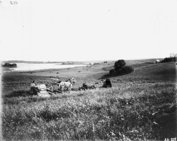 View down hill towards man using a horse-drawn McCormick binder in a field. In front of the binder is a man in a horse-drawn carriage. In the background is a large body of water.