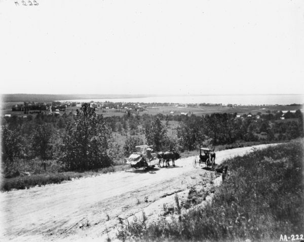 View down hill towards a man on a McCormick binder on a transport truck. Nearby is a horse-drawn carriage. In the far background is a town and a large body of water.