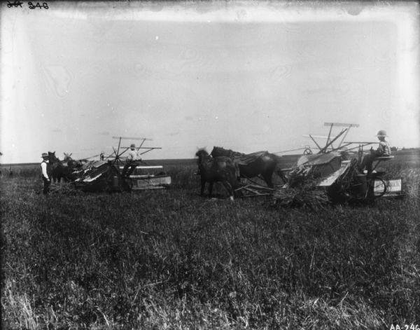 View across section of harvested field towards two people, one a young boy, using horse-drawn McCormick binders. A man is standing off to the left watching.
