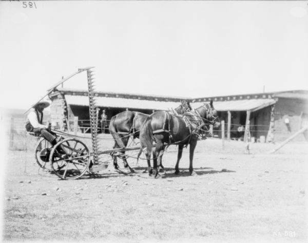 Right side view of a man sitting on a horse-drawn mower. The blade is in the upright position. In the background is a building with a roof over the open porch. One man is standing under the porch on the left, and a man is sitting under the porch roof on the right.