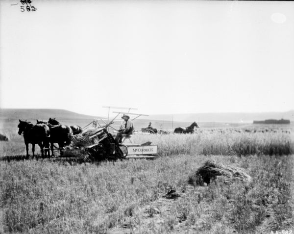 View across section of harvested field towards a man posing on a horse-drawn McCormick binder pulled by three horses. Behind him is another man in a horse-drawn buggy. Low hills are in the far background.