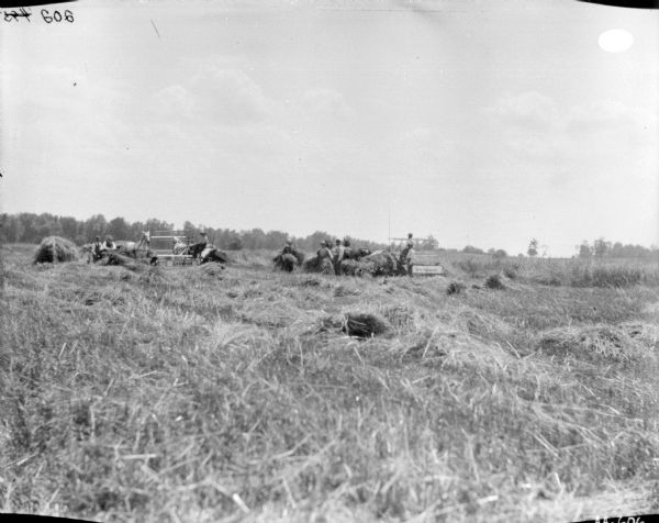View across field towards two men using horse-drawn McCormick binders in a field. Two men are in a carriage on the left near one of the binders. A group of four men are standing near the binder on the right.