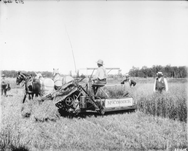 View from rear of a man using a horse-drawn McCormick binder in a field. A man is standing in front of the binder on the left. On the right, two men stand watching.