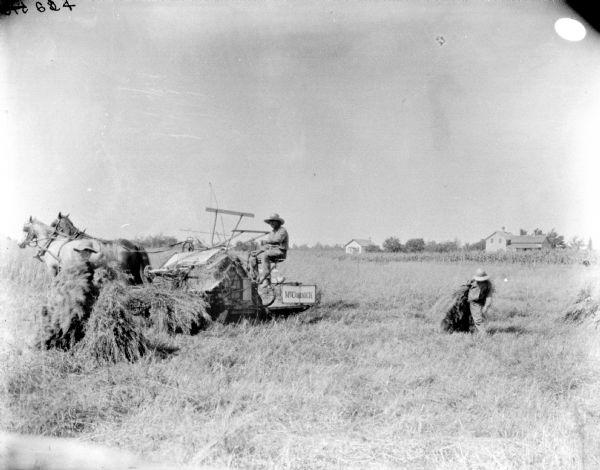 Man operating a horse-drawn McCormick combine in a field. Two young boys are working with sheaves of grain nearby. Farm buildings are in the background.