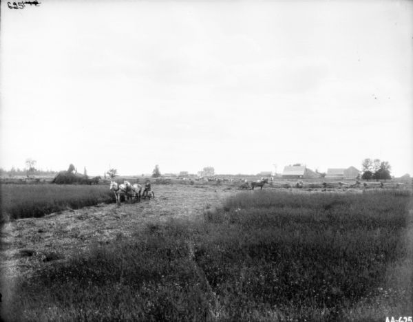 View across field towards a man on a horse-drawn mower. Behind him on the left men are working on and around a large haystack with horses. Behind a split-rail fence are cattle. A group of people are near the fence. In the far background are farmhouses and farm buildings.