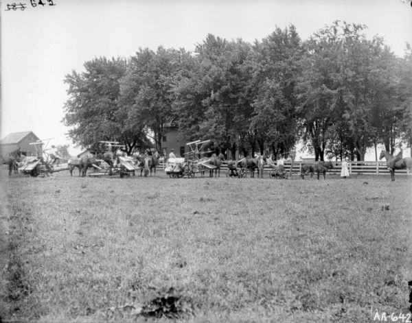 View across field towards group of people posing with three horse-drawn binders and two mowers. Men are sitting on the binders and mowres, and a group of men and women stand near the fence on the right. One man is sitting bareback on a horse on the far right. Behind the group is a fence, a farmhouse among trees, and other farm buildings.