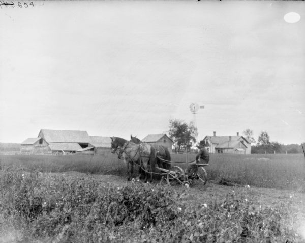 View from left rear of a man on a horse-drawn mower in a field. In the background are farm buildings and a windmill.
