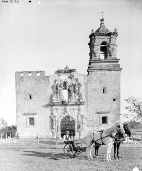 Man posing on a horse-drawn mower in front of the ruins of a building surrounded by a fence. The building is probably a church as there are wreaths on graves which are surrounded by the fence. The ruins have ornate details around the arch, windows and columns around the front entrance, and a bell tower is on the right.