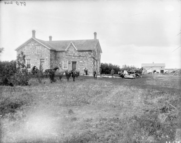 Group of people posing in front of a house. A man is sitting on a horse-drawn McCormick binder on the right, and in the background is a barn. One man is posing on a bicycle on the lawn, and a woman is standing near the house with a dog. Two women are sitting in a buggy on the left in front of the house.