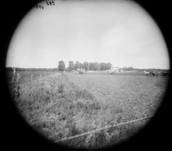 Circular-framed view along fence line of a field towards two or three men using horse-drawn mowers. A man is riding in a carriage or buggy on the far right. In the background are farm buildings.