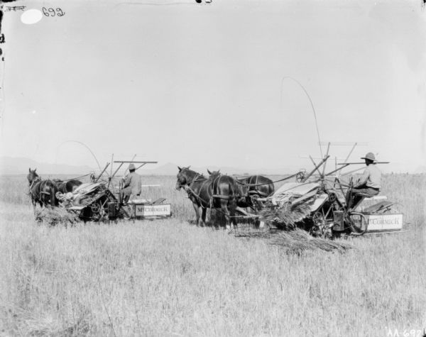 Three-quarter view from rear left of two men operating two horse-drawn McCormick binders in a field. Mountains are in the far background.