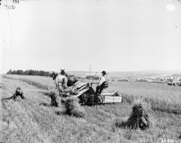 View from rear of a man using a horse-drawn McCormick binder in a field. Stacks of grain are in the field. In the background in a valley is a town.