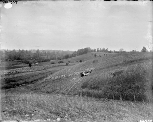 View down hill towards a man using a horse-drawn McCormick binder in a hilly field. Three men are standing on top of the hill on the right. On the left is a road through a valley.