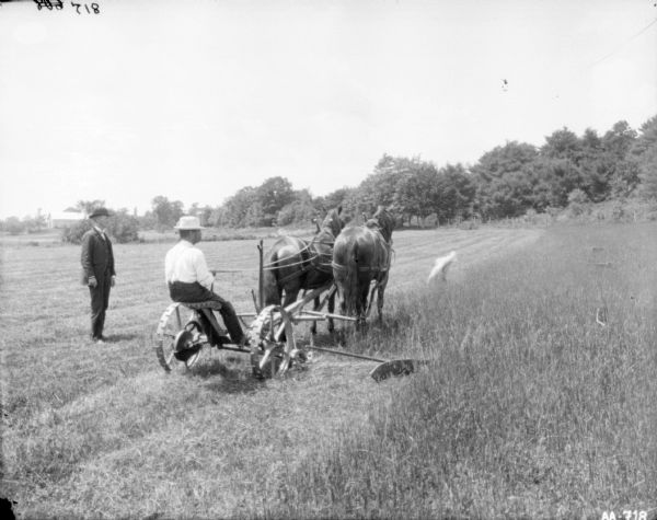 Three-quarter view from right rear of a man using a horse-drawn mower in a field. Another man, wearing a suit, is standing on the left in the mowed section of the field. Behind him in the background is a farm building among trees.