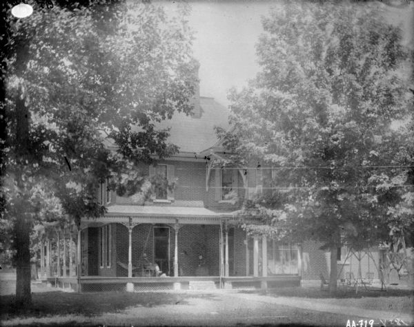 Family posing on the front porch of a large brick home. There is appears to be someone standing in a second-floor window above the porch. There is a lawn swing under trees on the right. Two people are standing on the far right side of the house, near the wood base of a windmill or water tower.