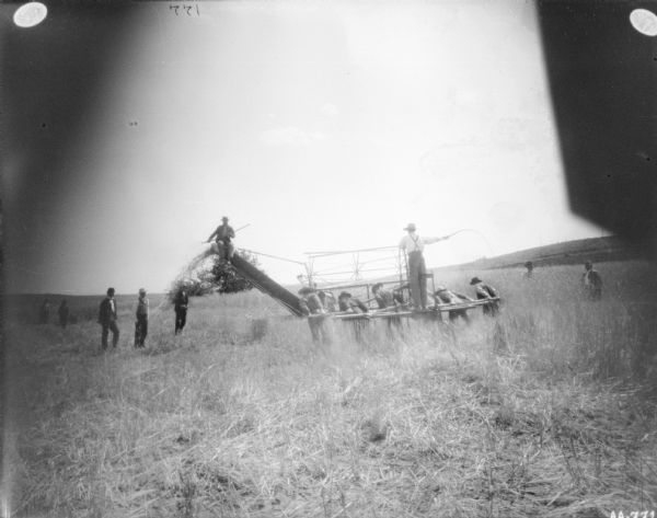 View from rear of six men pulling a binder across a field. A man is standing on the back of the binder holding a whip in one hand. Other men are standing and watching on the left and right. Another man is sitting high up on the top of a chute holding a rake.
