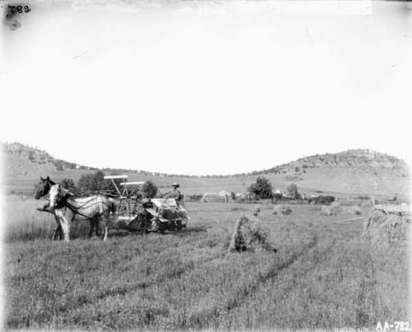 Man on horse-drawn binder in a partially harvested field. The horses are wearing fly-nets. A wagon is parked in the field on the far right. Behind him are farm buildings, and in the far background are rocky hills.