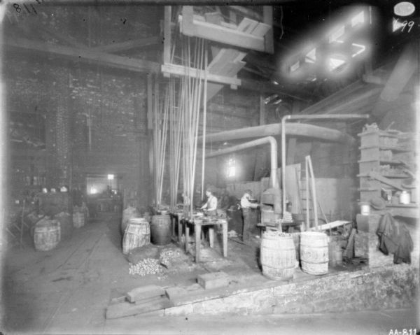 Men working in a  manufacturing area of the factory. Belt-driven machinery is attached to the high ceiling of the brick factory. Barrels are sitting on the floor. A small furnace is against the wall on the right.