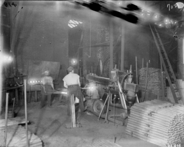 Men working in a manufacturing area at McCormick Works. The men are heating rods at a machine. On the floor beside the machine are more metal rods.