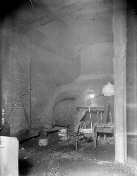 A man is standing on a wood bench to work at a furnace. The walls around the furnace are of brick.