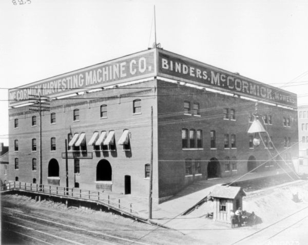 Slightly elevated view of dealership building. Railroad tracks are in the foreground, along two sides of the building, near the loading docks of the factory. Three men stand near a small building at the edge of the railroad tracks. There are brick archways along the sides of the factory near the loading docks.