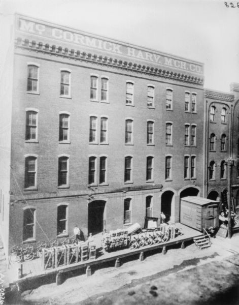Elevated view looking down towards the loading dock area of a dealership. There is a railroad car parked in the archway of the building on the right. There are men standing on the loading dock near agricultural machinery and a loaded cart.
