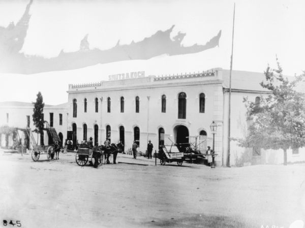 View from street of horse-drawn vehicles and men standing in front of the Smuts & Koch dealership. There is a McCormick binder at the corner of the building on the right.