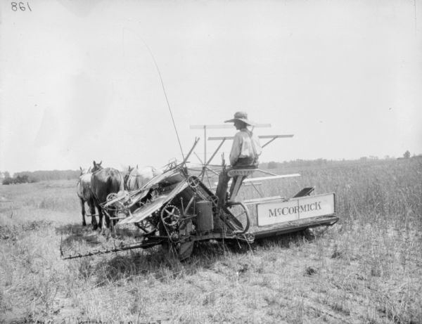 Three-quarter view from left rear of a man using a horse-drawn McCormick binder in a field.