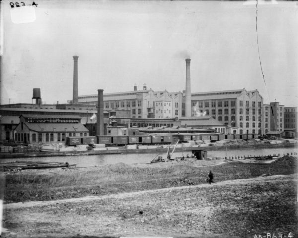 View across river towards the McCormick Works. Railroad cars are on railroad tracks near the factory buildings. Stacks of lumber are stacked in front of the railroad cars. A man is walking on a path in the foreground, and a group of men are on a boat at the near shoreline.
