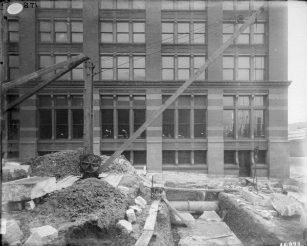Crane and building materials in the street in front of the McCormick Harvesting Machine Co. building at 212 Market Street. Two men are standing at the windows on the first floor of the building.