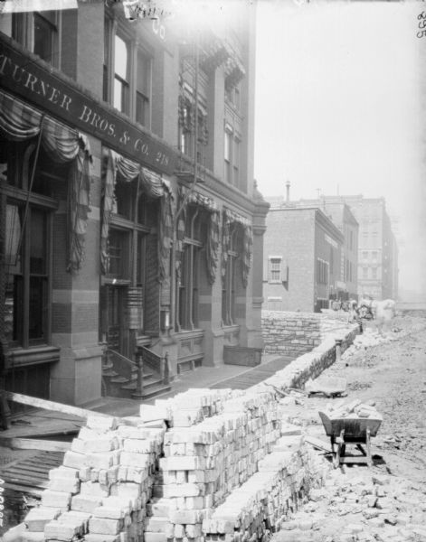 View of street being constructed outside the dealership office of the McCormick Harvesting Machine Co. on 212 Market Street. Stacks of cobblestones are in the foreground. There is a sign for "Turner Bros. & Co. 218" on the left. Further down the street men are working near a horse.