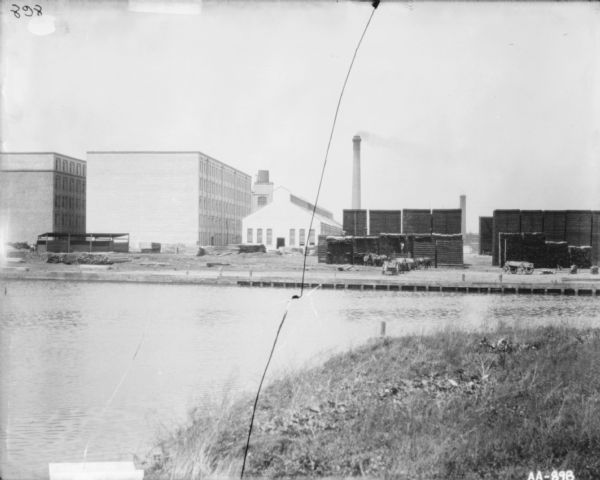 View from shoreline across river towards a yard with lumber at McCormick Works. Men are working in the yard near horses and wagons.